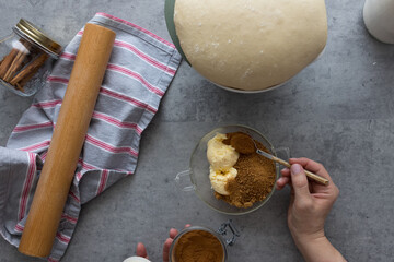 Woman hand pouring cinnamon powder with a spoon in a bowl full of panela and butter next to a risen dough.
