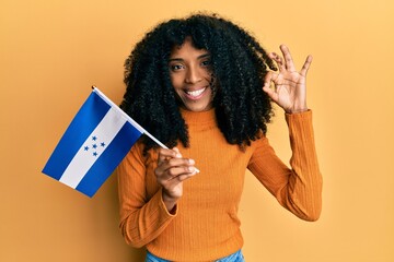 African american woman with afro hair holding honduras flag doing ok sign with fingers, smiling...
