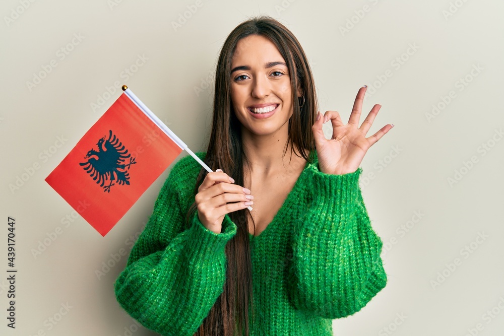 Wall mural young hispanic girl holding albania flag doing ok sign with fingers, smiling friendly gesturing exce