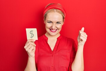 Young blonde woman holding dollar symbol reminder paper screaming proud, celebrating victory and...