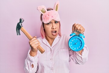 Hispanic woman with pink hair wearing pajama holding hammer and alarm clock in shock face, looking...