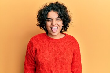 Young hispanic woman with curly hair wearing casual winter sweater sticking tongue out happy with funny expression. emotion concept.