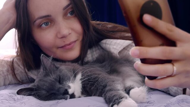 A charming woman smiles sweetly at the moment of taking pictures of herself with a fluffy domestic cat lying on the bed. The pet is sleeping. Selfie with animals on a smartphone.