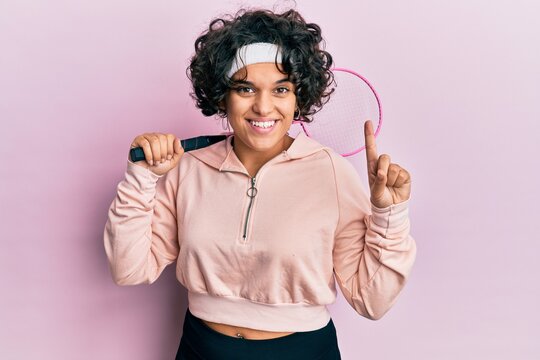 Young hispanic woman with curly hair holding badminton racket smiling with an idea or question pointing finger with happy face, number one