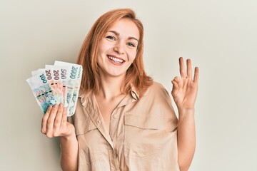 Young caucasian woman holding czech koruna banknotes doing ok sign with fingers, smiling friendly gesturing excellent symbol