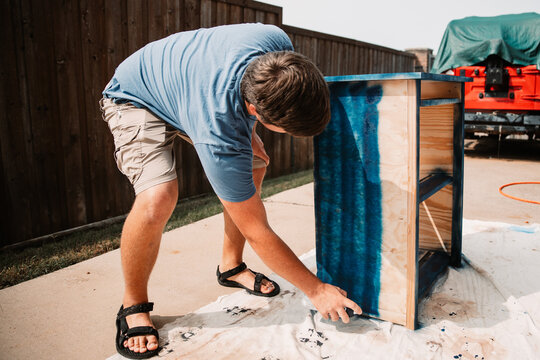 Young man spray paints an unfinished wooden furniture piece navy blue