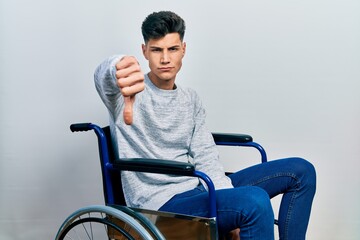 Young hispanic man sitting on wheelchair looking unhappy and angry showing rejection and negative with thumbs down gesture. bad expression.