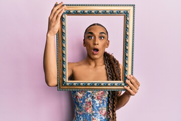 Hispanic man wearing make up and long hair holding vintage empty frame afraid and shocked with surprise and amazed expression, fear and excited face.