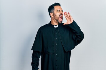 Handsome hispanic man with beard wearing catholic priest robe shouting and screaming loud to side...