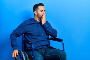 Handsome hispanic man with beard sitting on wheelchair bored yawning tired covering mouth with hand. restless and sleepiness.