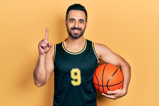 Handsome hispanic man with beard holding basketball ball surprised with an idea or question pointing finger with happy face, number one