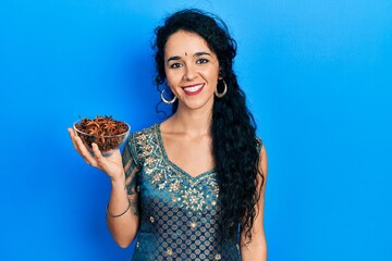 Young woman wearing bindi and traditional kurta dress holding bowl of star anise looking positive and happy standing and smiling with a confident smile showing teeth