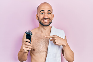 Young bald man shirtless holding electric razor smiling happy pointing with hand and finger