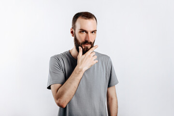 Serious puzzled handsome male model with beard holding hand on chin as if thinking about something, squinting at camera with suspicious look and standing over gray background. Man decides what to buy