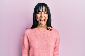 Young brunette woman with bangs wearing casual winter sweater sticking tongue out happy with funny...