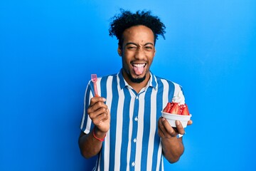 Young african american man with beard eating strawberry ice cream sticking tongue out happy with funny expression.