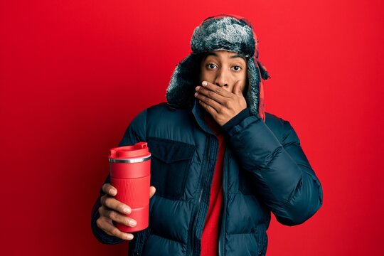 Young african american man with beard wearing winter hat holding hot coffee on thermo covering mouth with hand, shocked and afraid for mistake. surprised expression