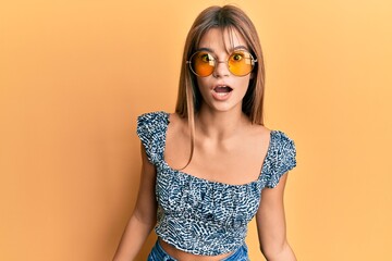 Teenager caucasian girl wearing fashion yellow sunglasses scared and amazed with open mouth for surprise, disbelief face