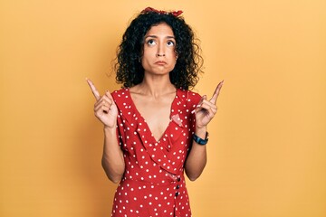 Young latin girl wearing summer dress pointing up looking sad and upset, indicating direction with fingers, unhappy and depressed.