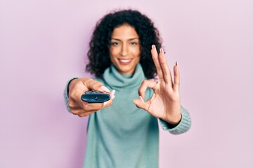 Young latin girl holding television remote control doing ok sign with fingers, smiling friendly gesturing excellent symbol
