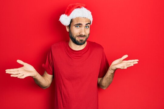 Young hispanic man wearing christmas hat clueless and confused expression with arms and hands raised. doubt concept.