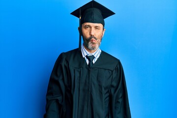 Middle age hispanic man wearing graduation cap and ceremony robe making fish face with lips, crazy...