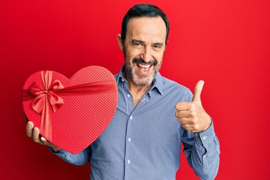 Middle age hispanic man holding valentine day gift smiling happy and positive, thumb up doing excellent and approval sign