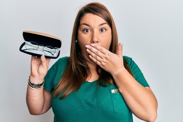 Beautiful brunette plus size woman holding glasses in eyewear case covering mouth with hand,...