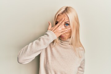 Young blonde woman wearing casual winter turtleneck sweater peeking in shock covering face and eyes with hand, looking through fingers afraid