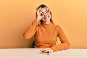 Young caucasian woman wearing casual clothes and glasses sitting on the table smiling happy doing ok sign with hand on eye looking through fingers