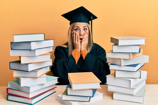 Young caucasian woman wearing graduation ceremony robe sitting on the table afraid and shocked, surprise and amazed expression with hands on face