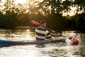 Active young caucasian guy looking concentrated while paddling kayak on a lake surrounded by nature...