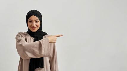 Smiling young arabic woman in hijab pointing to side - obrazy, fototapety, plakaty