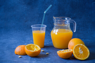 Naklejka na ściany i meble freshly squeezed orange juice in a jug and in a glass, next to halves of oranges and peel, orange still life on a blue background