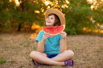 Adorable baby girl in straw hat sit with cross legs on green grass, and eagerly eats juicy...