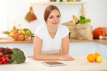 Obraz na płótnie Canvas An attractive young woman looking for a new recipe for a delicious salad mix while sitting at the table in sunny kitchen. Tablet pc is the best cookbook