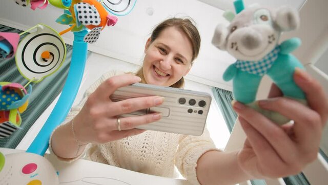 Smiling young mother shaking rattle toy to her baby and making photograph on smartphone for social media. Concept of parenting and making images of children