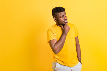 Pensive african american man in yellow t-shirt holding hand on chin looking at copy space, thoughtful doubtful black male with unsure face thinking,making decision isolated on yellow studio background