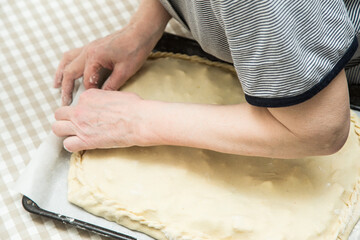 The hands of an adult elderly woman are preparing a dough pie. The fingers of the hands fasten the edges of the dough to each other.