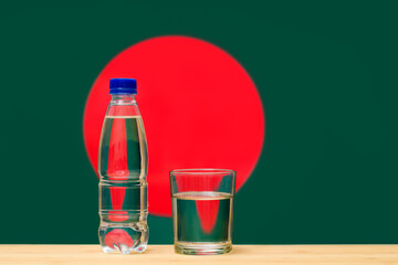 A bottle of clean drinking water and a glass stand on the table against the background of the flag of Bangladesh. A concept for the supply of clean drinking water in Bangladesh.