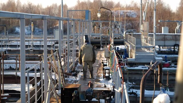 Worker with welding mask walks along bridge over reservoirs with dirty water at purification station in winter evening