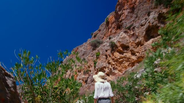 Safari Trip: Traveling Woman Walks in Zakros Gorge Rocks in Crete, Greece. Female Tourist in Hat Hikes in Mounting Enjoying Views and Trekking on Vacation in Summer. 4K Hand Held Tracking Wide Shot
