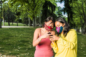 Obraz premium Lesbian latin lesbian couple texting with smartphone technology and face mask during covid-19 pandemic. young yellow and lavender. LGTBQ