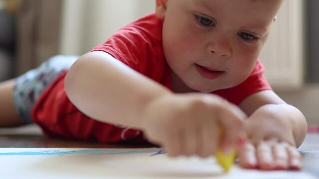 close-up view of portrait of a toddler in red shirt lying on the floor at home and drawing with crayons, early kids education concept
