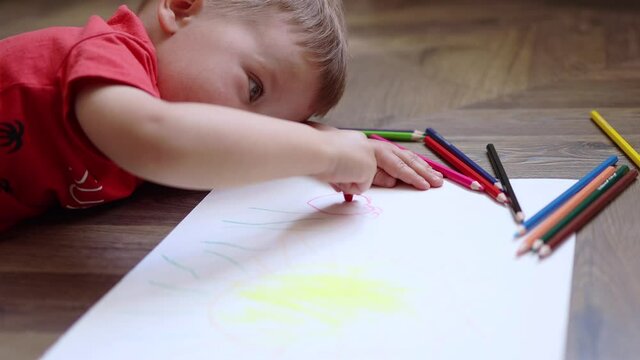 low angle view of little baby boy in red clothes lying on the floor and drawing summer picture with colorful pencils crayons, home education early learning concept