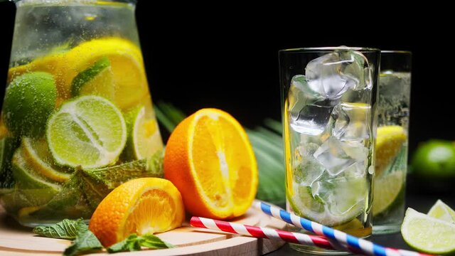 Close-up of citrus fruit drink in jug, glasses with ice and carbonated water, shooting of cocktail of orange, lemon and lime on black background, drinking cold lemonade with mint and sliced fruits. 