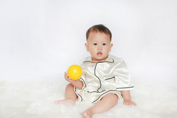 Portrait of little Asian baby boy in pajamas holding yellow plastic toy ball sitting on white fur against white background.