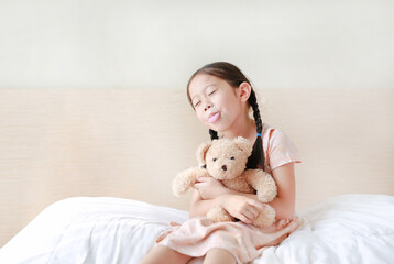 Funny of Asian little girl embracing teddy bear with tongue sticking out while sitting on the bed at home.