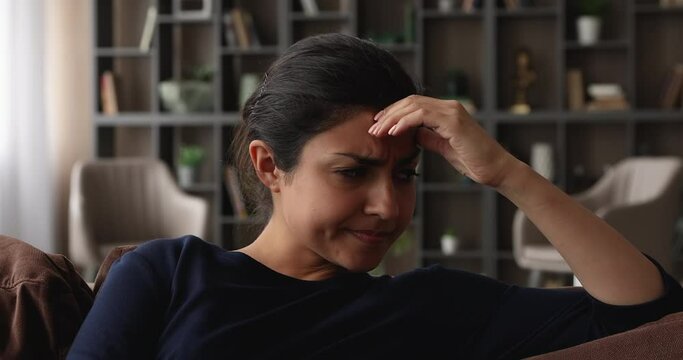 Close up face of Indian woman sit on sofa deep in sad thoughts feels resentment, angry with yourself because of mistake, frowning looking upset. Break up, bad news, unpleasant life situation concept