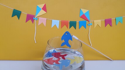 Kites, flags, colorful fish in the jar with water, fishing rod, on the table. Yellow background. Tradition to brighten up the Brazilian June party is the play and fun of toy fishing. Space for text. - obrazy, fototapety, plakaty